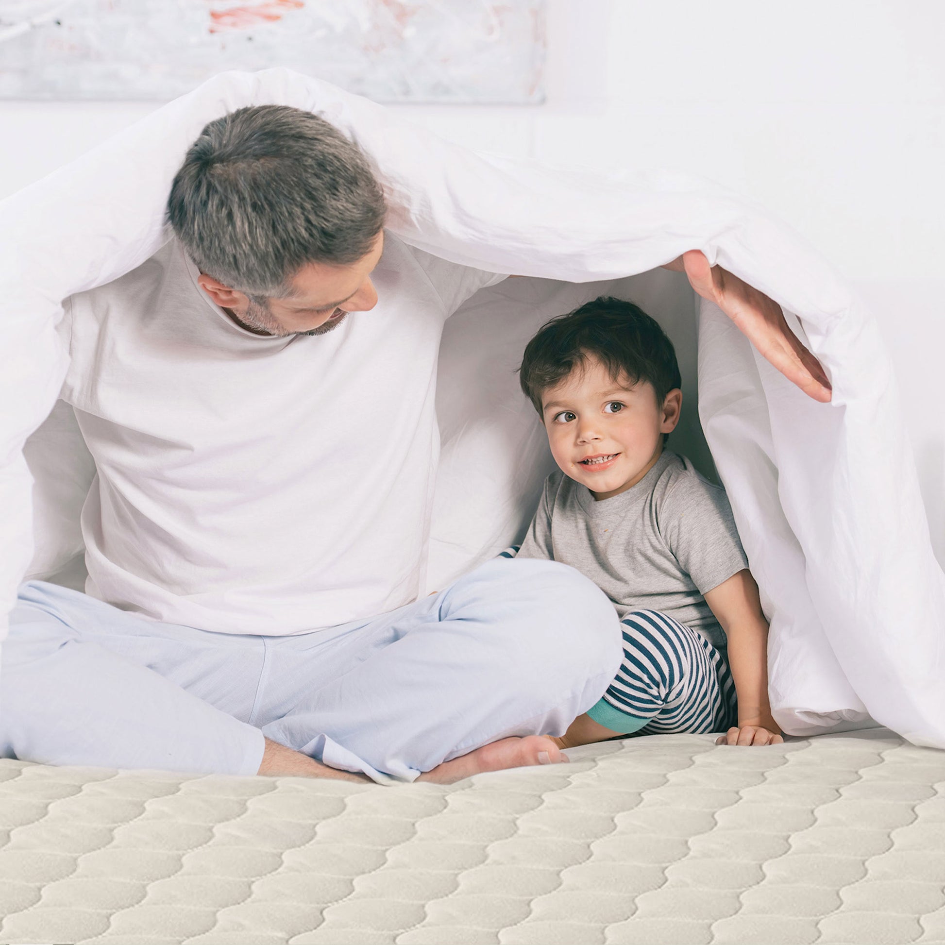 Father and son smiling on bed with organic cotton mattress pad under comforter.